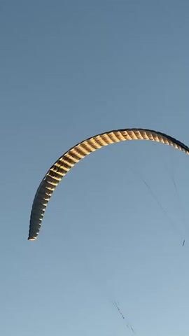 The video shows a kite in the sky during the day. The kite is curved and appears to be white and grey. It is attached to thin lines that extend downwards. The sky is clear and blue. The video then pans down to a beach scene at sunset. There are people on the beach, some structures, and a person appears to be holding onto the kite lines. The sun is setting on the right side of the frame, casting an orange glow.