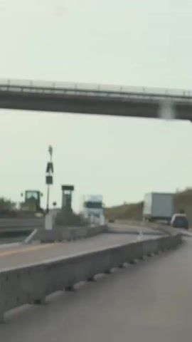 The video shows a highway scene with several vehicles. In the foreground, a concrete barrier lines the right side of the road. The road itself is paved, with white lane markings. A white semi-truck with the DAF logo is prominently featured in the center of the frame, driving towards the viewer. To the left of the truck, a dark-colored van is also visible. Further back, another white semi-truck with a trailer is seen on the right side of the road. In the far background, a lighter-colored vehicle is partially visible on the right. Above the road, a concrete overpass or bridge structure spans the scene. On the left side of the road, near the barrier, there are traffic signals with solar panels. The sky is overcast and appears bright. The overall environment suggests a road with moderate traffic.