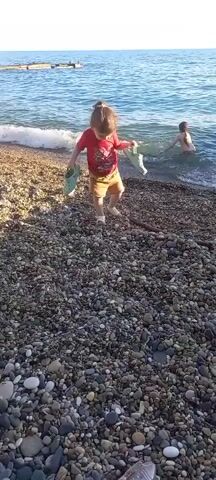 The video shows a young child standing on a pebble beach near the water. The child is wearing a red t-shirt and yellow shorts and is holding a light-colored object in each hand. The child appears to be looking at the water. In the background, another person is partially submerged in the ocean. The water is calm with small waves lapping at the shore. The sun appears to be shining, suggesting it is daytime. The scene is a beach, likely in a coastal area, with the ocean visible. The overall mood seems peaceful and relaxed. There is no visible text in the video.