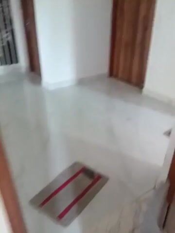 The video shows a panning view of an indoor environment with white marble tile flooring. The camera moves from a doorway with a wooden door, past two doormats, one closer and one further away, and into a larger space. The space appears to be a hallway or entryway, with white walls and a few wooden doors visible in the background. Light reflects off the polished floor, creating bright spots. In the foreground, a wooden chair or stool is partially visible. The overall impression is of a clean and modern interior.