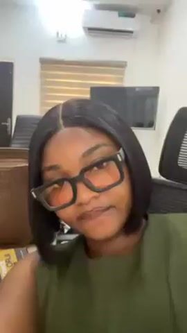 A woman with dark hair, wearing glasses and an olive green sleeveless top, is looking directly at the camera. She is indoors in an office-like environment. There are chairs, a desk, and a television visible in the background. The lighting in the room is bright. The camera is positioned at eye level. She appears to be speaking.