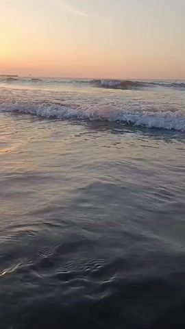 A first-person perspective video shows a beach at sunset. The camera is low to the ground, close to the water's edge. Gentle waves roll onto a dark, sandy shore. The sun is low in the sky, casting a warm, orange glow on the water and clouds. The sky is a gradient of orange and pink, with a few wispy clouds. In the distance, a faint structure, possibly a pier or a building, can be seen on the horizon. The sound of the ocean waves is present throughout the video.