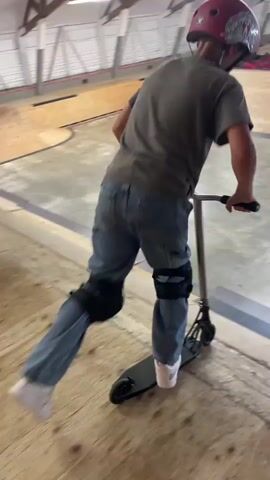 A person wearing a red helmet, gray shirt, denim jeans, knee pads, and white shoes is riding a scooter in an indoor skatepark. The person is on a ramp and then jumps over a gap. The skatepark has wooden ramps and walls. Other people are visible in the background sitting on ledges.
