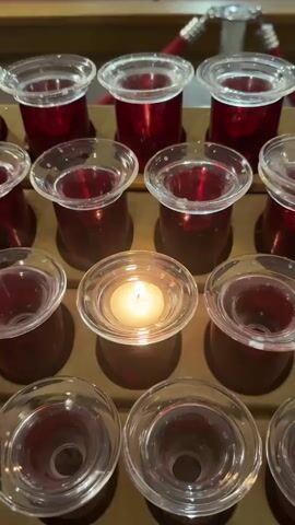 The video begins with a close-up of several votive candles in a church, one of which is lit. The camera then slowly pans up and back, revealing rows of wooden pews and the altar area of the church. A person in dark clothing is standing near the altar, facing away from the camera. The lighting is dim, creating a solemn atmosphere. The church appears to be empty except for the person at the altar.