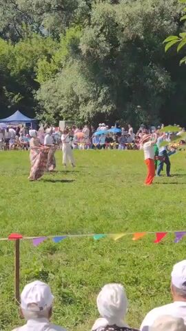 A group of people in colorful traditional clothing are participating in an outdoor activity on a grassy field. Some are holding objects that appear to be water guns, and water is being sprayed. There is a crowd of spectators in the background, some of whom are seated. The event is taking place on a sunny day, with trees and tents visible in the distance. Colorful flags are strung across a foreground area, suggesting a festive or celebratory atmosphere.
