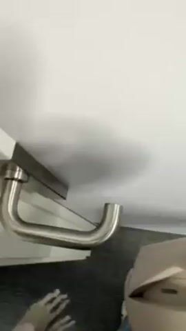The video shows a hand inserting a white card into a slot on a door handle. The door handle is silver and curved. The door and wall appear to be white. The floor is carpeted in a dark color. Part of a beige object, possibly a piece of luggage, is visible in the bottom right corner. The hand wears a sweater with a black and white woven pattern.