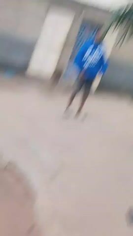 A man wearing a blue hoodie and shorts stands in a courtyard. He is facing the camera and appears to be posing. The courtyard has a dirt floor and is surrounded by buildings. There is a palm tree to the right of the man. Several objects are scattered on the ground, including what appear to be shoes and a metal basket. The lighting suggests it is daytime.