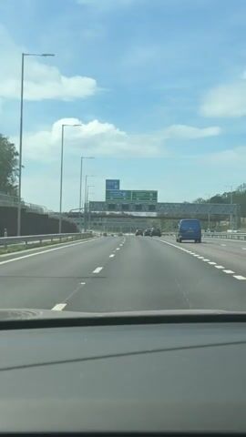 A first-person perspective video shows a car driving on a highway under a partly cloudy sky. The road ahead is relatively clear with a few other vehicles visible in the distance. Overhead signs indicate directions towards Gatwick, Heathrow, Portsmouth, and other locations via M25 and M23. There are road markings and street lights lining the highway. A "PASSED" sticker is visible on the windshield.