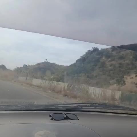 A car is driving on an asphalt road between two hills. The hills are covered with trees, dry grass, and shrubs. The sky is overcast. There are sunglasses on the car's dashboard. A motorcycle goes by in the other direction.