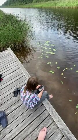 A child sits on a wooden dock with their feet dangling in the water of a narrow canal. The child is holding a phone in their hands and appears to be looking at it. There are several pairs of flip-flops on the dock next to the child. The canal is lined with tall green grass and trees. The water has lily pads floating on the surface. The sky is overcast.