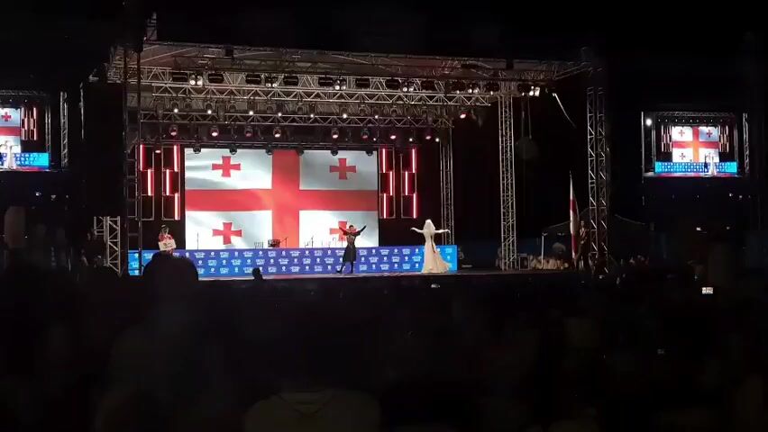 This video captures an outdoor nighttime event with a stage setup. The stage is illuminated with bright lights and features a large screen displaying the Georgian flag multiple times. Two figures are visible on the stage, one dressed in dark clothing and another in a long, light-colored garment. They appear to be performing or presenting. The stage is supported by a metal truss system, and additional screens are mounted on either side, also displaying the Georgian flag. In front of the stage, a crowd of people are gathered, silhouetted against the lights, suggesting they are spectators. The overall atmosphere seems festive, possibly a cultural festival or concert.