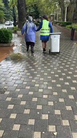 Two women are walking on a paved sidewalk. The woman on the left is wearing a blue tunic and a headscarf and is sweeping the sidewalk with a broom. The woman on the right is wearing a blue tunic, a high-visibility vest, and a headscarf, and is pushing a trash can on wheels. The sidewalk is wet, and there are scattered leaves on the ground. Trees and bushes line the sidewalk, and a street is visible in the background.