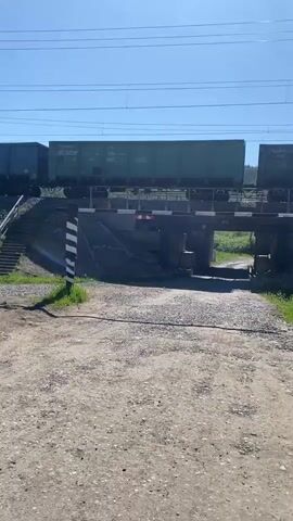A freight train crosses a railway bridge on a sunny day. The train consists of several hopper cars, some of which have Cyrillic lettering visible. The train is moving from left to right across the frame. Below the bridge, a set of stairs leads up to the tracks. A black and white striped pole stands in the foreground, likely part of a railway crossing signal. The ground in the foreground is gravelly. The sky is clear and blue.