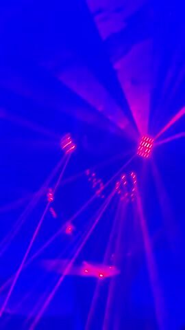 A person wearing a light-up suit is playing a drum set surrounded by purple and pink laser lights. The person's face is obscured by the lights and the angle of the shot. The drum set is partially visible. The environment is dark and appears to be a performance venue or club.