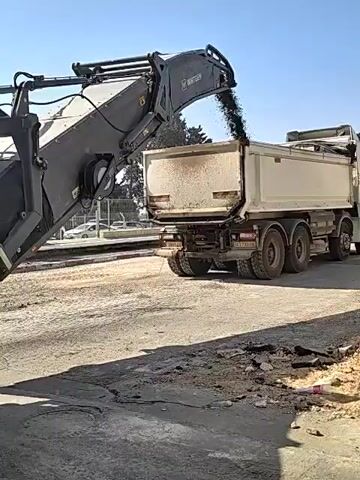 A Wirtgen milling machine is shown removing asphalt from a road surface. The removed asphalt is being loaded into the back of a dump truck. The scene appears to be a road construction or repair site. The sky is clear and blue. There are some trees and buildings visible in the background. The milling machine has a large drum with teeth that grinds away the asphalt. The asphalt is then conveyed up a ramp and deposited into the truck.