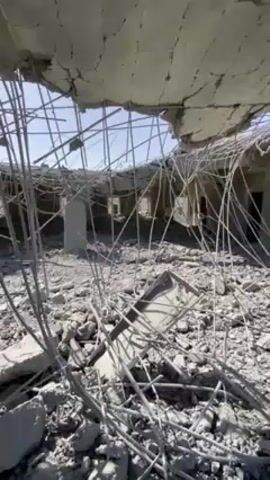 The camera pans across the interior of a destroyed building. The ceiling has partially collapsed, exposing twisted metal and rubble. What remains of the structure reveals empty rooms with dark doorways. The ground is covered in debris. In the distance, we can see a hint of a clear sky. The scene evokes a sense of devastation and ruin.