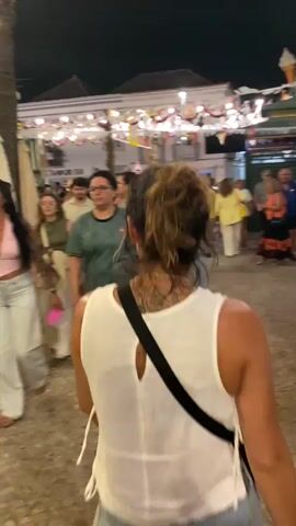 The video shows a street scene at night, likely a festival or outdoor market. The foreground is dominated by the back of a woman with her hair tied up, wearing a white tank top and denim shorts, with a black strap crossing her shoulder. She is walking away from the camera. To her left, a woman wearing a green and white soccer jersey and shorts walks in the same direction. To her right and further back, a group of people are visible, including a man in a blue shirt and shorts, and a woman in an orange top. The background features buildings decorated with lights and colorful banners or flags. The ground is paved with cobblestones. The overall impression is of a lively, crowded evening event.
