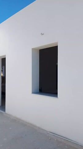 The video shows a slow pan of a corner of a white building on a clear, sunny day. The building has a flat roof and three door openings that lead into dark rooms. One wall also has a square window opening. The floor is concrete. There are some small objects on the roof, including what appears to be a piece of wood. The clear blue sky is visible above the building.