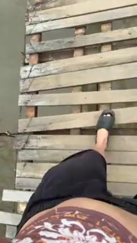 The video shows a person walking barefoot across a rickety wooden suspension bridge over a river. The person is carrying two bags, one in each hand, and is wearing a black t-shirt and brown shorts.  The bridge is made of rough-hewn wooden planks and supported by metal cables. The river below is flowing rapidly and is murky.  In the background, there is a grassy area with trees and buildings.  The sky is overcast and gray. The camera is in motion, following the person as they walk across the bridge.
