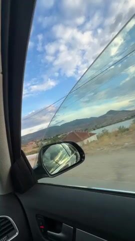 The video is filmed from inside a car, looking out the passenger side window. The window is partially open, and the side mirror is visible. The scene outside is a landscape with mountains in the distance, a body of water, and a small white building with a red roof. The sky is filled with dramatic clouds. The camera angle is tilted, giving the impression of the car moving. There are no people visible in the video, and no specific actions are taking place beyond the implied movement of the car. The overall impression is one of a scenic drive through a rural or mountainous area.