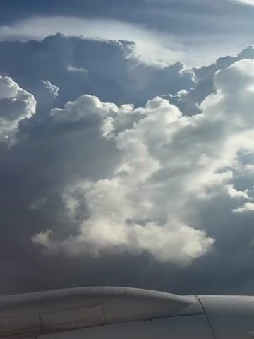 The video shows a view from an airplane window, looking out at a dramatic sky filled with large, textured clouds. The clouds are a mix of bright white where they are lit by sunlight and dark gray in the shadows, suggesting depth and scale. Some clouds appear to be towering cumulonimbus formations. The lower portion of the frame shows the metallic curve of an airplane engine nacelle, its surface reflecting some light. The sky above the clouds is a deep blue. The light suggests it is either early morning or late afternoon. There are no people visible.