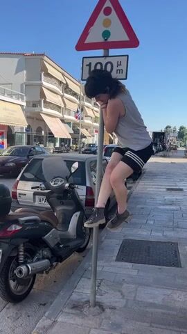 A young person with dark hair, wearing a grey tank top and black athletic shorts, is performing a pole dance routine around a metal pole in an urban environment. The pole is located on a sidewalk next to a street. Above the pole, there is a triangular traffic sign depicting a red, yellow, and green circle, indicating a traffic light ahead, and a rectangular sign below it with the number "100" followed by Greek letters. The person is holding onto the pole with their arms and legs, demonstrating flexibility and strength as they move their body in a fluid motion. Their legs are extended, one bent at the knee and the other straight, wrapped around the pole. The person's body is contorted into various positions as they ascend and descend the pole. In the background, there are parked cars, buildings with shops on the ground floor, and a scooter parked to the left of the pole. The sun is shining, casting shadows on the ground, suggesting it is a bright, sunny day. The pavement is made of interlocking stone tiles, and there is a metal manhole cover visible on the right side of the image. The person's movements are graceful and controlled, indicating practice and skill in this form of performance.