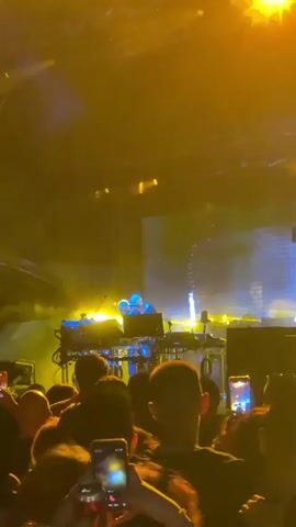 The video shows a person on a stage behind a console, with bright lights and smoke. The person appears to be a DJ or musician. The stage is lit with yellow and white lights. A large screen behind the person displays a pixelated image. In the foreground, there is a crowd of people watching the performance. Some of the people in the crowd are holding up their phones to record the event. The overall atmosphere is one of a live music concert.