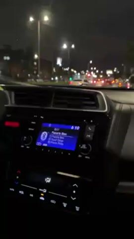 A car navigates a city street at night, with light posts illuminating the road and the city lights adding ambient light to the dark sky. The car's dashboard is visible, showing a digital display with music information and controls. In the foreground, the car's interior appears dark, with the dashboard buttons and controls visible. Outside, traffic moves along the street with headlights shining. The scene captures the feeling of a late-night drive through an urban environment.