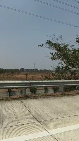 The video shows a view from a moving vehicle. The foreground consists of a concrete road surface with white lines marking lanes. A metal guardrail runs along the side of the road, separating it from an area with dry grass and some vegetation. Beyond the guardrail, there are trees with green leaves and a landscape with open fields. In the distance, there are tall poles with wires stretching across the sky, likely power lines. The sky is clear and appears to be a bright, sunny day. The overall environment suggests an outdoor, possibly rural or semi-rural, setting.