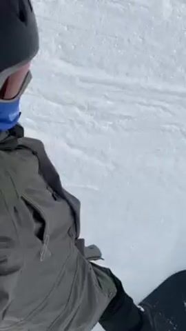 A snowboarder navigates down a snowy slope. The person is wearing a grey and black jacket with a fur-lined hood, a black helmet, ski goggles, and a blue neck gaiter. The snowboarder is moving downhill, with snow visible all around. Red poles and a safety net are seen on the side of the slope. Trees are visible in the background.