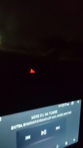 The video shows a nighttime drive on a highway. The perspective is from inside a vehicle, looking forward. The road is visible with lane markings and reflective markers along the side. There are a few other vehicles on the road, including trucks, whose headlights and taillights are illuminated. A blue overhead highway sign is visible in the distance. The sky is dark, indicating it is nighttime. There are a few small, blurry objects hanging from the top of the windshield, possibly decorative items. The dashboard of the vehicle is partially visible at the bottom of the frame, showing a red indicator light and a screen displaying what appears to be a time of 8:17 AM and some text in a non-English language. The lighting conditions are low, with the primary light sources being the vehicle headlights, taillights, and road markers. The footage appears to be slightly shaky, consistent with being recorded while driving.