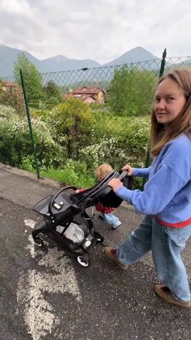Here's a description of the video:

A young woman wearing a blue sweater and jeans pushes a black stroller along a paved path. Seated in the stroller is a toddler with blond hair, dressed in an orange jacket. The stroller has a grey seat and a black frame with four wheels.

The woman pushes the stroller forward, with the toddler looking towards a green chain-link fence that runs parallel to the path. Behind the fence is a hedge with white flowers, and in the background, there are houses and mountains.

The camera focuses on the toddler, who is holding onto the stroller's handlebar. As the stroller moves, the background blurs, indicating motion.