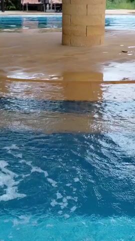 The video opens with a close-up of blue water in a swimming pool. The camera then pans to a pillar made of tan bricks, with a sandy colored surface surrounding it. The surface is wet and appears to be a poolside area. In the background, there is a swimming pool with lounge chairs and umbrellas. A building with a dark roof and balconies is visible behind the pool. The scene suggests a tropical or resort-like setting.