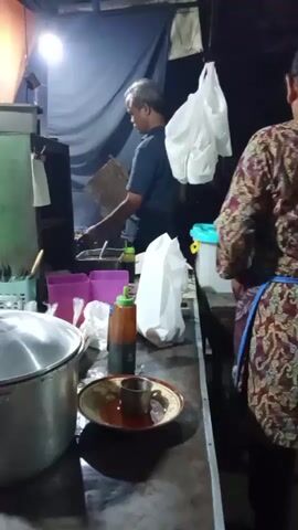 A man is cooking at a food stall at night. He is wearing a dark t-shirt and has gray hair. He is standing behind a counter with various cooking utensils and ingredients. A woman in a patterned dress is standing to the right of the frame, also behind the counter. She is wearing an apron. There are plastic bags hanging above the counter, and various containers and bottles on the counter. The lighting is dim and appears to be from a single overhead light source. The overall environment suggests a busy street food vendor.