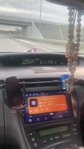 The media file shows the interior of a car, specifically the dashboard and windshield. The car is in motion, driving on a highway with multiple lanes. The time displayed on the car's dashboard is 19:45, indicating it's late evening. The weather appears cloudy.

Prominently displayed on the dashboard is a car's infotainment system, showing a music player interface. The song playing is titled "mal-chishnik.noch-dj.kolya.remix". Other text on the screen includes "Неизвести" (Unknown), "ЧАСТОТА" (Frequency), and "Нет LRC файлов" (No LRC files). A phone is mounted in a holder to the left of the infotainment screen.

Hanging from the rearview mirror is a beaded string with a crucifix at the end. The overall mood is one of travel, with the focus on the car's interior and the activity of driving, accompanied by music. The highway appears to be in a semi-urban or rural setting, with some greenery visible beyond the road. The presence of other vehicles on the highway suggests it's a moderately trafficked route.