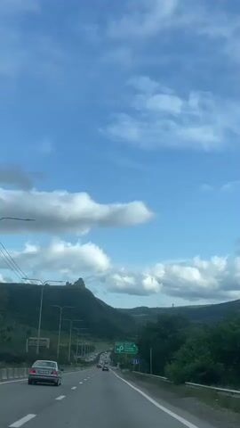 A car travels down a highway on a sunny day. The sky is blue with puffy white clouds.  Mountains covered in green trees rise on either side of the road. Other cars are visible on the highway in the distance. The car passes a sign with green lettering and a picture of a parking symbol. A guardrail runs along the side of the road, and the camera appears to be shooting from inside the car. The view is partially obscured by the car’s mirror.
