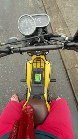 This video is a first-person perspective shot from a motorcycle. The rider is wearing pink pants and pink Crocs. The motorcycle is yellow and has a brown seat. The handlebars are black and have a carbon fiber wrap. The dashboard has a speedometer and an odometer. The road is wet, indicating it has recently rained. In the background, there are trees, a metal guardrail, and a sign that says "PHONG NHA - KE BANG" on a mountain. The sky is overcast.