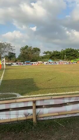 A wide shot shows a grassy sports field with a white line running across it. In the background, there are tents, people, and trees. The sky is cloudy.