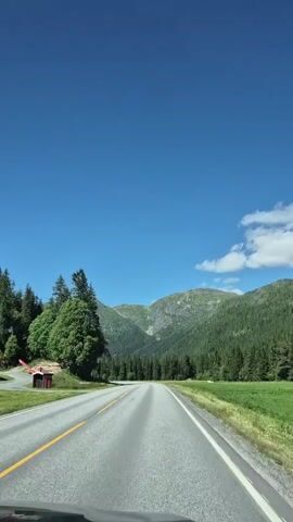 The video shows a car driving on a paved road through a landscape of mountains and trees. The sky is blue with a few white clouds. The road is asphalt with yellow and white lines. On the left side of the road, there are trees and a small red building. On the right side, there is a green field. The mountains in the background are covered with trees. The car is moving forward and the scenery is passing by.