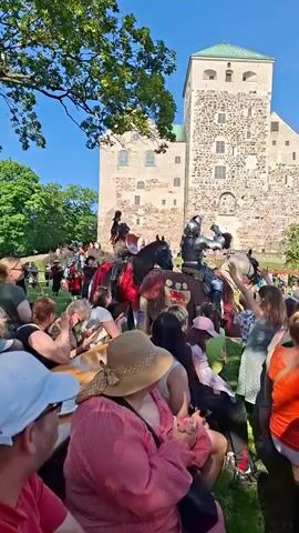 This is a short, vertical video clip, likely filmed at a medieval reenactment event. The scene is dominated by two knights in full armor, mounted on black horses adorned with red and gold caparisons. One knight on the left has a shield with a red heart and a gold border. The other knight on the right has a helmet with a beak-like protrusion and a shield. Both knights are holding lances, suggesting they are preparing for or engaged in a joust. Behind them, a stone castle with multiple windows stands under a clear blue sky. In the foreground and surrounding the knights, a crowd of spectators is visible, many seated on the grass, some standing, and several people in the front rows are looking towards the knights. The overall atmosphere appears festive and historical.