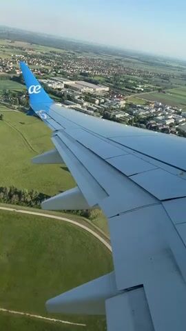 The view from an airplane window shows the wing of the plane with the "ae" logo on the tail. Below, a green field, a walking path, and buildings that appear to be a water park with multiple pools can be seen. Beyond that is a town with houses, trees, and fields. The sky is blue.