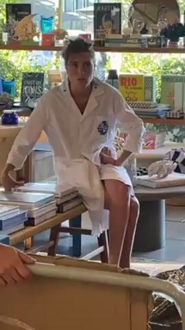 A woman wearing a white lab coat sits on a bench next to a table filled with books and magazines. The background includes a window with foliage and a shelf displaying various decorative items and books. Another person is partially visible in the foreground, holding a metal frame. The woman looks directly at the camera, speaks, and smiles broadly.