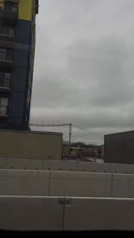 The video shows a moving train arriving at the Bryn Mawr station. The weather is overcast and the surrounding area includes buildings, trees, and a billboard. A person is standing on the platform. The Bryn Mawr station sign is visible.