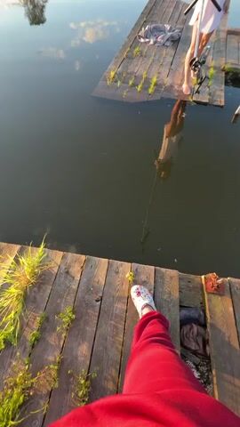 A first-person perspective shows a person's legs in red pants and a floral shoe standing on a wooden dock. In front of them, on the water, a woman in a white dress stands on a wooden raft, holding a long pole with a fishing net attached. The woman is attempting to catch something in the water with the net, and the person on the dock appears to be assisting or encouraging her. The sun is setting, casting a warm glow on the scene, and the water reflects the sky and clouds.