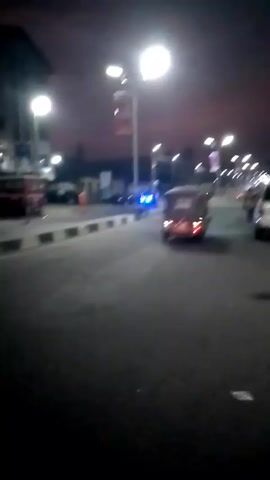 The video shows a street at night, likely in an urban area. Streetlights illuminate the scene, and the sky has a faint reddish glow. Several vehicles are visible, including cars and auto-rickshaws (tuk-tuks), driving along the road. A pedestrian walks near a parked white SUV on the right side of the street. There are poles with flags or banners along the roadside. The overall scene is somewhat blurry.