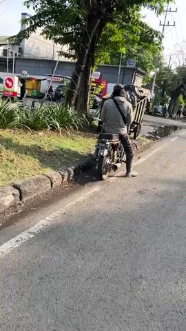 This media file captures a street scene in Sumorame, East Java. The primary subject is a person sitting on a motorcycle, facing away from the camera. They are wearing dark pants, a gray hooded jacket, and a dark cap. A backpack or bag is slung over their shoulder. The motorcycle appears to be an older model with a license plate that reads "L 5868 FYD".

Behind the motorcycle, a cart-like structure is visible, possibly a mobile vendor's cart or a makeshift trailer, with some bags or items on it. Further in the background, there are buildings, trees, and other vehicles, including a white car and a motorcycle. A red and white sign with the word "KELUAR" (meaning "Exit" in Indonesian) is visible on the left. Another sign further back seems to read "PURIS".

The scene appears to be taking place outdoors, possibly on the side of a road or a street. The lighting suggests it is either early morning or late afternoon, with the sun casting long shadows. The ground is paved, and there's a curb with grass and some foliage alongside it. There are puddles of water on the road, indicating recent rain or damp conditions. The overall atmosphere seems casual and ordinary, a typical street scene in a local area. The weather appears to be calm.
