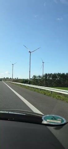 A video captures a view from inside a car driving on a road. In the distance, a line of three wind turbines stands against a clear blue sky. The turbines are tall, white structures with three blades each, and they appear to be in operation, with their blades slowly rotating. A guardrail runs along the side of the road, separating it from a grassy area dotted with trees. The road itself is paved and marked with white lines. The interior of the car is visible in the foreground, with a portion of the dashboard and windshield in view. A sticker with a green and white emblem is affixed to the dashboard. The overall scene suggests a daytime drive through a rural landscape with renewable energy infrastructure.