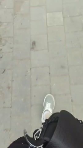 The video begins with a close-up shot of a person's legs and feet as they walk on a paved sidewalk. One person is wearing a black skirt and grey sneakers with white socks. A bandage is visible on their right leg. The camera then pans up to show a view of a park or recreational area. There are trees, grass, and a paved path. In the distance, a body of water, possibly a lake or river, is visible, with hills or mountains in the background. A playground with swings and other equipment can be seen near the water, and several people are present in the playground area. The sky is overcast. A wooden pole stands prominently in the foreground for a portion of the video.
