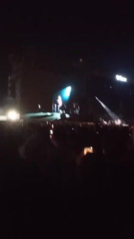 The video shows a concert at night. A large stage is set up with a performer on it, highlighted by a spotlight. The stage also has large screens displaying visuals. A crowd of people is visible in the foreground, many holding up their phones to record the performance. Lights flash and move across the stage, creating an energetic atmosphere. The video is shaky, indicating the camera operator is part of the crowd and possibly moving or dancing.