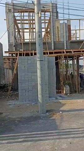 The video shows a construction site with a building under construction. The building appears to be two stories high and is partially completed. There are stacks of cinder blocks, scaffolding made of bamboo, and piles of gravel visible. In the background, there are other houses and a flag pole with a flag. A motorcycle is seen riding past the construction site. The overall environment appears to be a developing residential area.